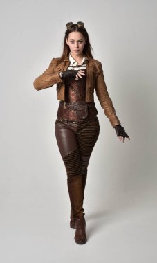 tam uzunlukta kahverengi deri steampunk kıyafeti giymiş esmer kız portresi. gri studio arka plan üzerinde ayakta poz.