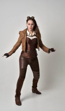 tam uzunlukta kahverengi deri steampunk kıyafeti giymiş esmer kız portresi. gri studio arka plan üzerinde ayakta poz.