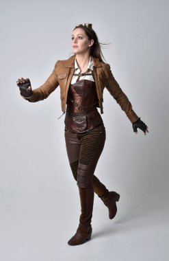 tam uzunlukta kahverengi deri steampunk kıyafeti giymiş esmer kız portresi. gri studio arka plan üzerinde ayakta poz.