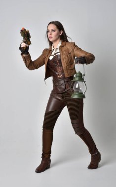 tam uzunlukta kahverengi deri steampunk kıyafeti giymiş esmer kız portresi. elinde bir silah, gri studio arka plan üzerinde ayakta poz.