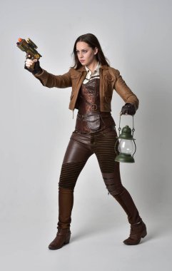 tam uzunlukta kahverengi deri steampunk kıyafeti giymiş esmer kız portresi. elinde bir silah, gri studio arka plan üzerinde ayakta poz.