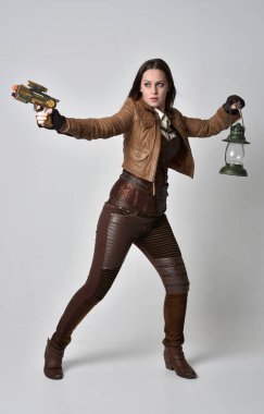tam uzunlukta kahverengi deri steampunk kıyafeti giymiş esmer kız portresi. elinde bir silah, gri studio arka plan üzerinde ayakta poz.