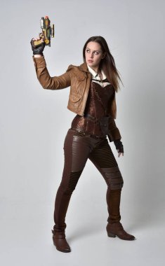 tam uzunlukta kahverengi deri steampunk kıyafeti giymiş esmer kız portresi. elinde bir silah, gri studio arka plan üzerinde ayakta poz.