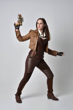 tam uzunlukta kahverengi deri steampunk kıyafeti giymiş esmer kız portresi. elinde bir silah, gri studio arka plan üzerinde ayakta poz.