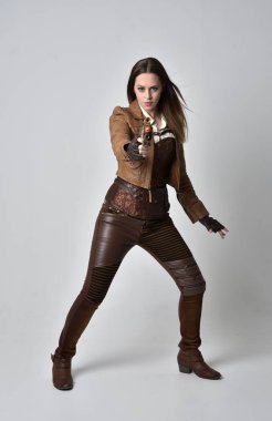 tam uzunlukta kahverengi deri steampunk kıyafeti giymiş esmer kız portresi. elinde bir silah, gri studio arka plan üzerinde ayakta poz.
