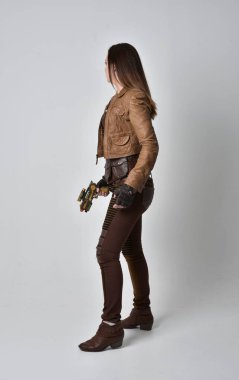 tam uzunlukta kahverengi deri steampunk kıyafeti giymiş esmer kız portresi. elinde bir silah, gri studio arka plan üzerinde ayakta poz.
