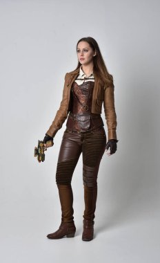 tam uzunlukta kahverengi deri steampunk kıyafeti giymiş esmer kız portresi. elinde bir silah, gri studio arka plan üzerinde ayakta poz.