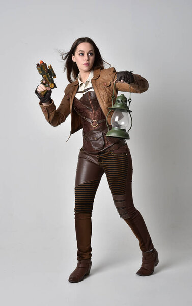 full length portrait of brunette  girl wearing brown leather steampunk outfit. standing pose holding a gun, on grey studio background.