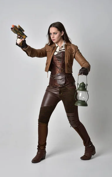 stock image full length portrait of brunette  girl wearing brown leather steampunk outfit. standing pose holding a gun, on grey studio background.