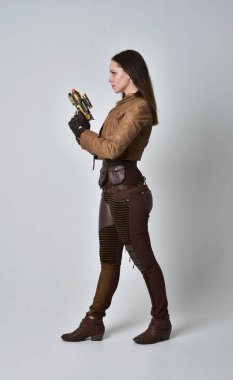 tam uzunlukta kahverengi deri steampunk kıyafeti giymiş esmer kız portresi. elinde bir silah, gri studio arka plan üzerinde ayakta poz.