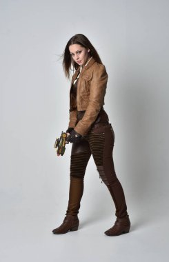 tam uzunlukta kahverengi deri steampunk kıyafeti giymiş esmer kız portresi. elinde bir silah, gri studio arka plan üzerinde ayakta poz.