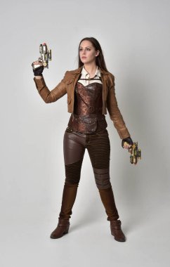 tam uzunlukta kahverengi deri steampunk kıyafeti giymiş esmer kız portresi. elinde bir silah, gri studio arka plan üzerinde ayakta poz.