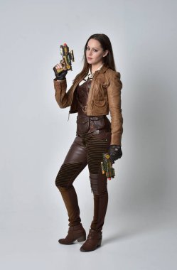 tam uzunlukta kahverengi deri steampunk kıyafeti giymiş esmer kız portresi. elinde bir silah, gri studio arka plan üzerinde ayakta poz.