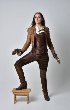 full length portrait of brunette  girl wearing brown leather steampunk outfit. standing pose holding a gun, on grey studio background. clipart