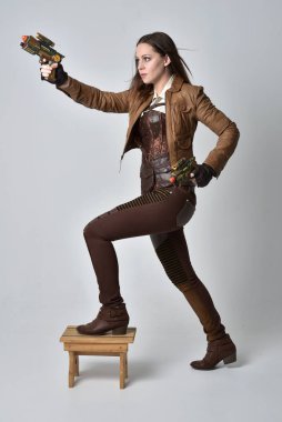 tam uzunlukta kahverengi deri steampunk kıyafeti giymiş esmer kız portresi. elinde bir silah, gri studio arka plan üzerinde ayakta poz.