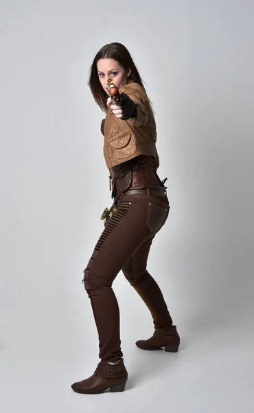穿棕色皮革蒸汽朋克服装的黑发女孩的全长肖像 站在姿势拿着枪 在灰色的工作室背景 — 图库照片