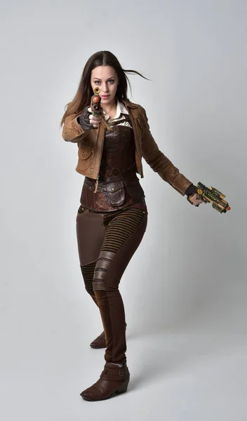 穿棕色皮革蒸汽朋克服装的黑发女孩的全长肖像 站在姿势拿着枪 在灰色的工作室背景 — 图库照片