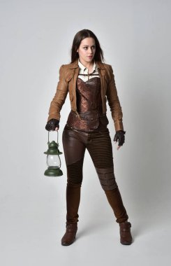 tam uzunlukta kahverengi deri steampunk kıyafeti giymiş esmer kız portresi. bir gaz fener gri studio arka planda tutarak ayakta poz.