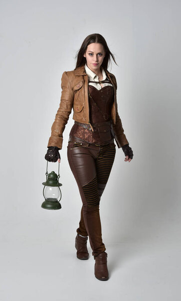 full length portrait of brunette  girl wearing brown leather steampunk outfit. standing pose holding a gas lantern on grey studio background.