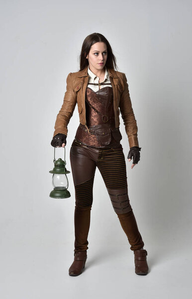 full length portrait of brunette  girl wearing brown leather steampunk outfit. standing pose holding a gas lantern on grey studio background.