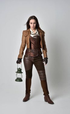 tam uzunlukta kahverengi deri steampunk kıyafeti giymiş esmer kız portresi. bir gaz fener gri studio arka planda tutarak ayakta poz.