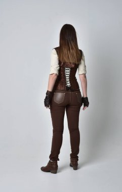 tam uzunlukta kahverengi deri steampunk kıyafeti giymiş esmer kız portresi. ayakta poz ile kameranın arkasında gri studio arka plan.