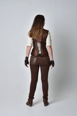 tam uzunlukta kahverengi deri steampunk kıyafeti giymiş esmer kız portresi. ayakta poz ile kameranın arkasında gri studio arka plan.