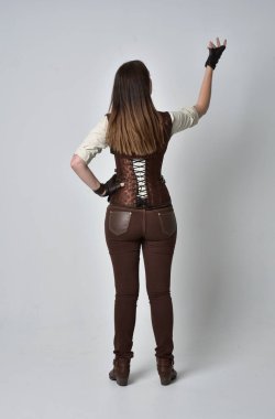 tam uzunlukta kahverengi deri steampunk kıyafeti giymiş esmer kız portresi. ayakta poz ile kameranın arkasında gri studio arka plan.