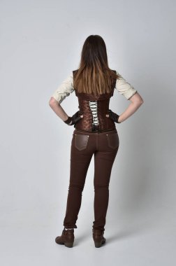tam uzunlukta kahverengi deri steampunk kıyafeti giymiş esmer kız portresi. ayakta poz ile kameranın arkasında gri studio arka plan.