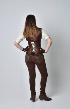 tam uzunlukta kahverengi deri steampunk kıyafeti giymiş esmer kız portresi. ayakta poz ile kameranın arkasında gri studio arka plan.