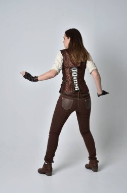 tam uzunlukta kahverengi deri steampunk kıyafeti giymiş esmer kız portresi. ayakta poz ile kameranın arkasında gri studio arka plan.