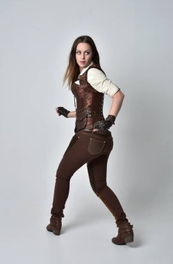 tam uzunlukta kahverengi deri steampunk kıyafeti giymiş esmer kız portresi. ayakta poz ile kameranın arkasında gri studio arka plan.