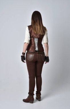 tam uzunlukta kahverengi deri steampunk kıyafeti giymiş esmer kız portresi. ayakta poz ile kameranın arkasında gri studio arka plan.