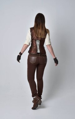 tam uzunlukta kahverengi deri steampunk kıyafeti giymiş esmer kız portresi. ayakta poz ile kameranın arkasında gri studio arka plan.