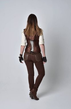 tam uzunlukta kahverengi deri steampunk kıyafeti giymiş esmer kız portresi. ayakta poz ile kameranın arkasında gri studio arka plan.