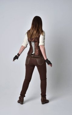 tam uzunlukta kahverengi deri steampunk kıyafeti giymiş esmer kız portresi. ayakta poz ile kameranın arkasında gri studio arka plan.