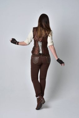 tam uzunlukta kahverengi deri steampunk kıyafeti giymiş esmer kız portresi. ayakta poz ile kameranın arkasında gri studio arka plan.