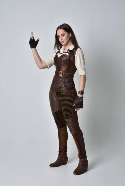 tam uzunlukta kahverengi deri steampunk kıyafeti giymiş esmer kız portresi. gri studio arka plan üzerinde ayakta poz.