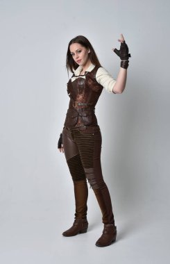 tam uzunlukta kahverengi deri steampunk kıyafeti giymiş esmer kız portresi. gri studio arka plan üzerinde ayakta poz.