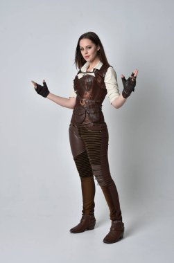 tam uzunlukta kahverengi deri steampunk kıyafeti giymiş esmer kız portresi. gri studio arka plan üzerinde ayakta poz.