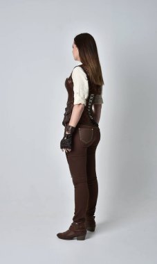 tam uzunlukta kahverengi deri steampunk kıyafeti giymiş esmer kız portresi. gri studio arka plan üzerinde ayakta poz.