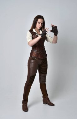 tam uzunlukta kahverengi deri steampunk kıyafeti giymiş esmer kız portresi. gri studio arka plan üzerinde ayakta poz.