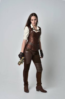 tam uzunlukta kahverengi deri steampunk kıyafeti giymiş genç esmer kız portresi. elinde bir silah gri studio arka plan üzerinde ayakta poz.