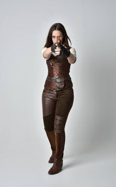 full length portrait of young, brunette  girl wearing brown leather steampunk outfit. standing pose holding a gun on grey studio background.