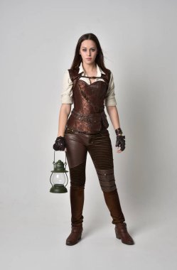 tam uzunlukta kahverengi deri steampunk kıyafeti giymiş genç esmer kız portresi. bir gaz fener gri studio arka planda tutarken ayakta poz.