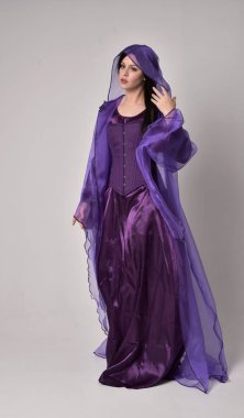 tam uzunlukta mor fantezi Ortaçağ elbisesi ve pelerin giymiş uzun siyah saçlı güzel kız portresi. gri studio arka plan üzerinde ayakta poz.