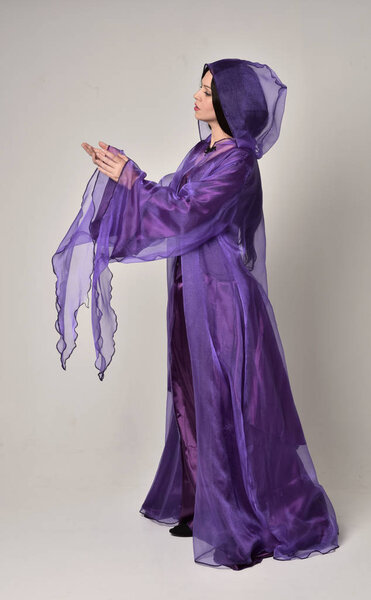 full length portrait of beautiful girl with long black hair,   wearing purple fantasy medieval gown and cloak. standing pose on grey studio background.