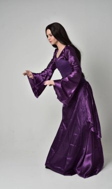 tam uzunlukta portresi uzun siyah saçlı güzel kız mor fantezi Ortaçağ elbisesi giyiyor. gri studio arka plan üzerinde ayakta poz.