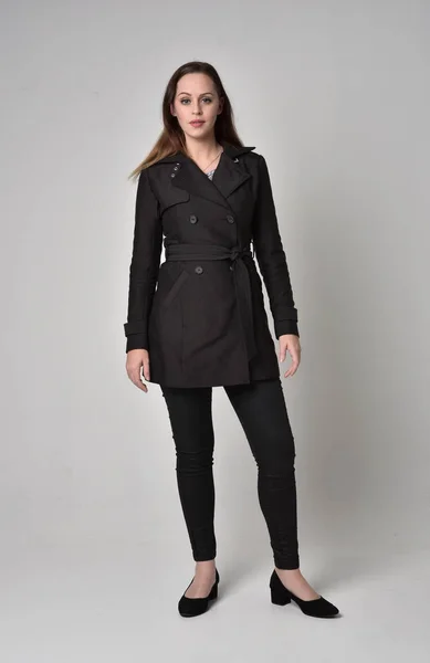 一个穿黑色长外套的黑发女孩的全长肖像 站在灰色的工作室背景摆姿势 — 图库照片