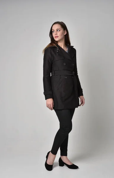 stock image full length portrait of a brunette girl wearing long black coat, standing pose on grey studio background.